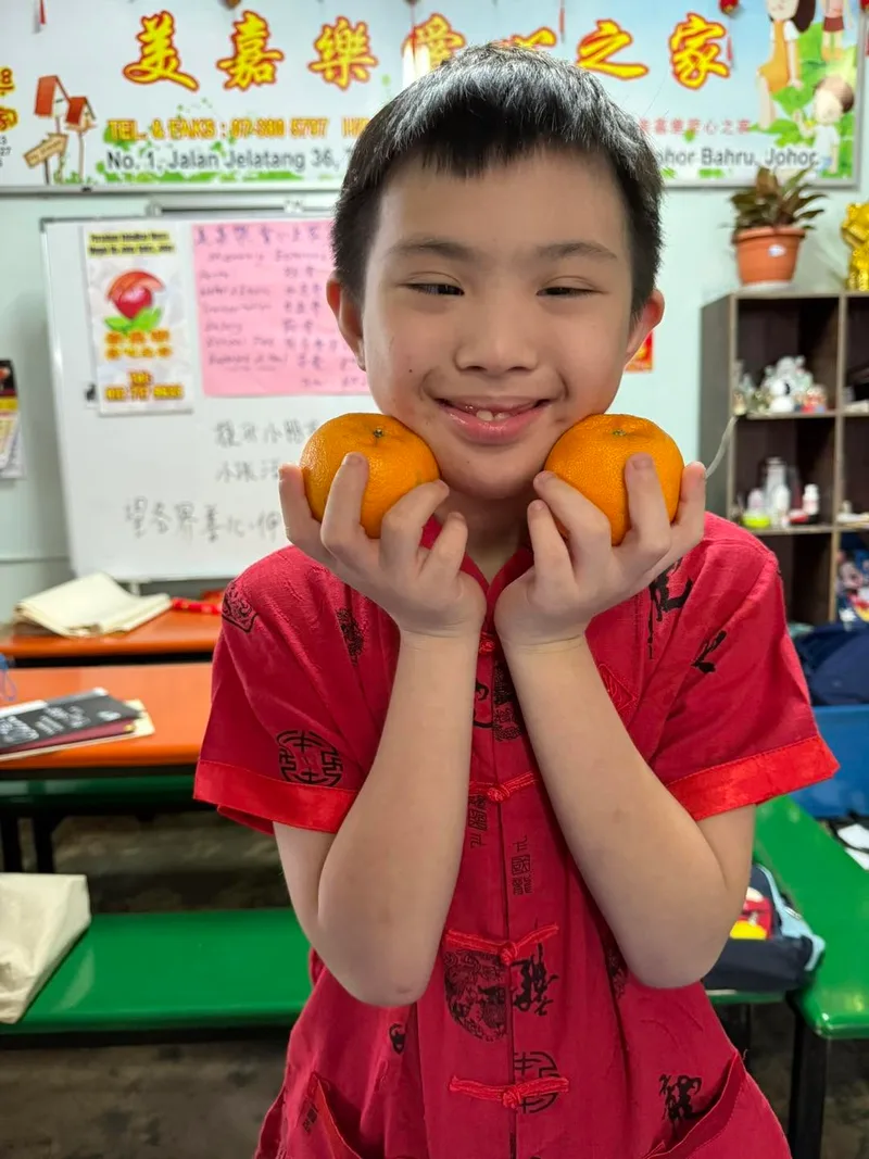 A smiling boy in a red festive shirt holding a mandarin orange to each cheek