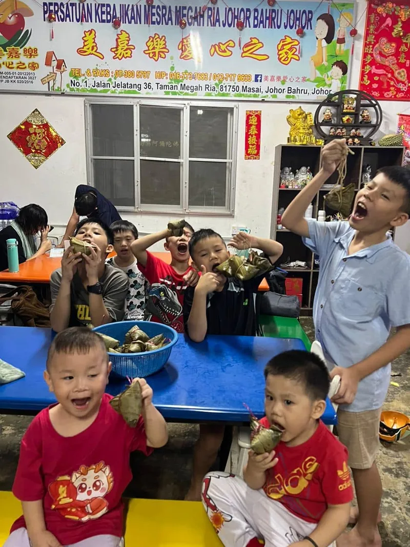 A group of boys at a table eating zongzi rice dumplings, pulling playful open-mouthed faces, inside the shelter's common room