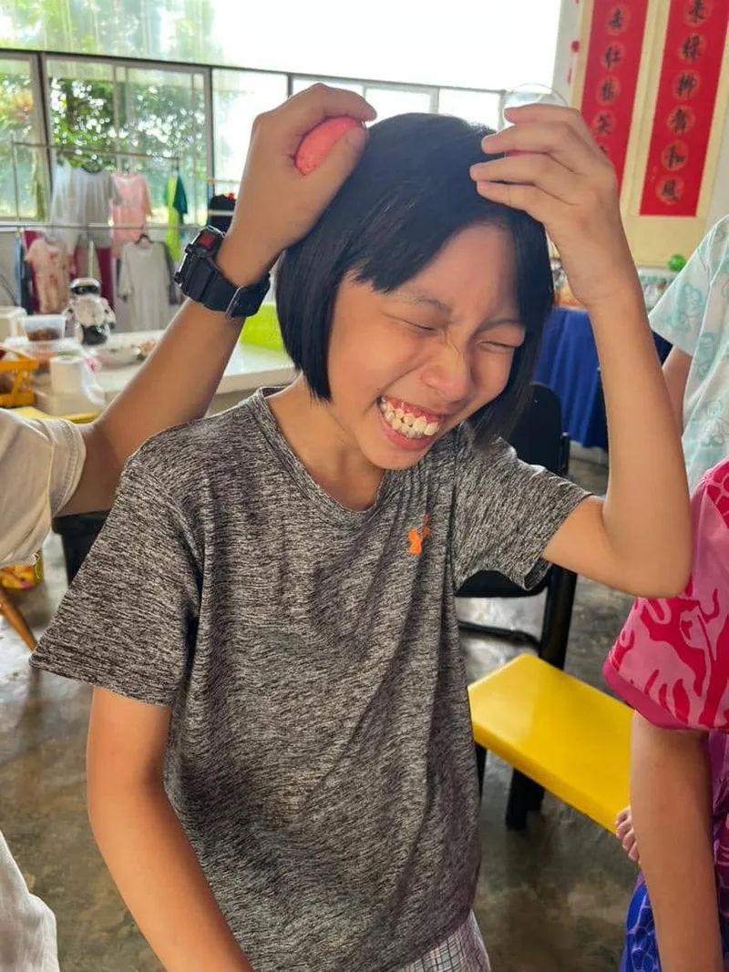 A girl in a grey T-shirt laughing with her eyes closed, hands raised to her head, red Chinese New Year couplets on the wall behind her