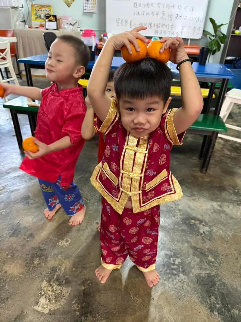 A toddler in a red and gold festive outfit balancing two mandarin oranges on his head, another boy in red beside him