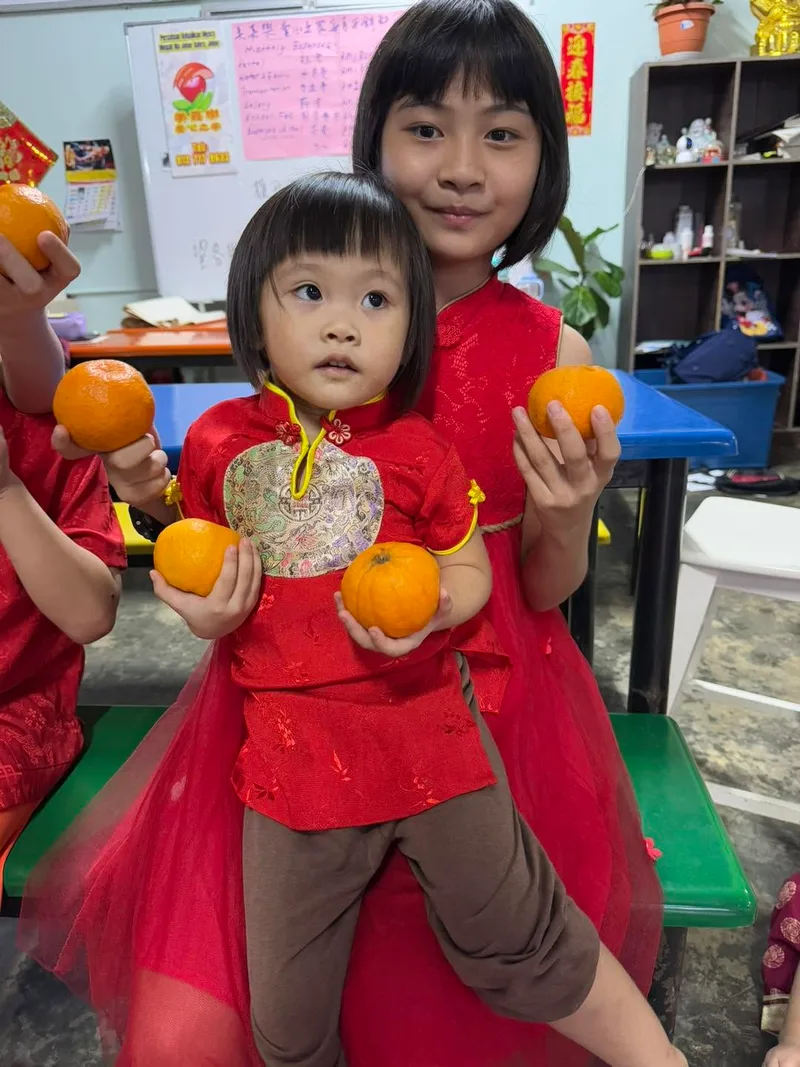 Two girls in red dresses, an older girl with her arm around a younger girl, both holding mandarin oranges